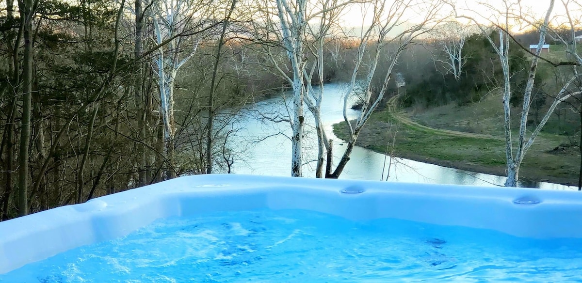 A hot tub is positioned on a deck, offering a view of the Shenandoah River below. The scene captures the water’s gentle flow and the surrounding trees, reflecting soft evening light. The tranquil setting invites relaxation amidst natural beauty.