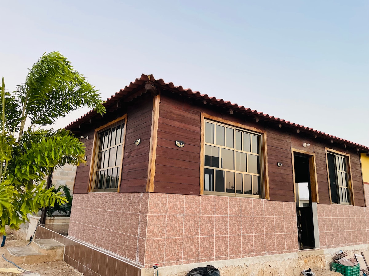 The exterior of the house features a combination of wooden accents and tiled walls. Large windows allow natural light to fill the interior, showcasing a welcoming design. A palm tree stands nearby, enhancing the serene environment.