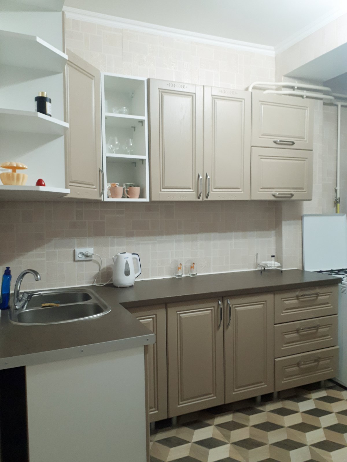 The kitchen area features light-colored cabinetry with upper shelves holding glassware. A stainless steel sink is situated on a countertop, accompanied by an electric kettle. An oven is visible in the corner, with patterned flooring adding a subtle detail to the space.