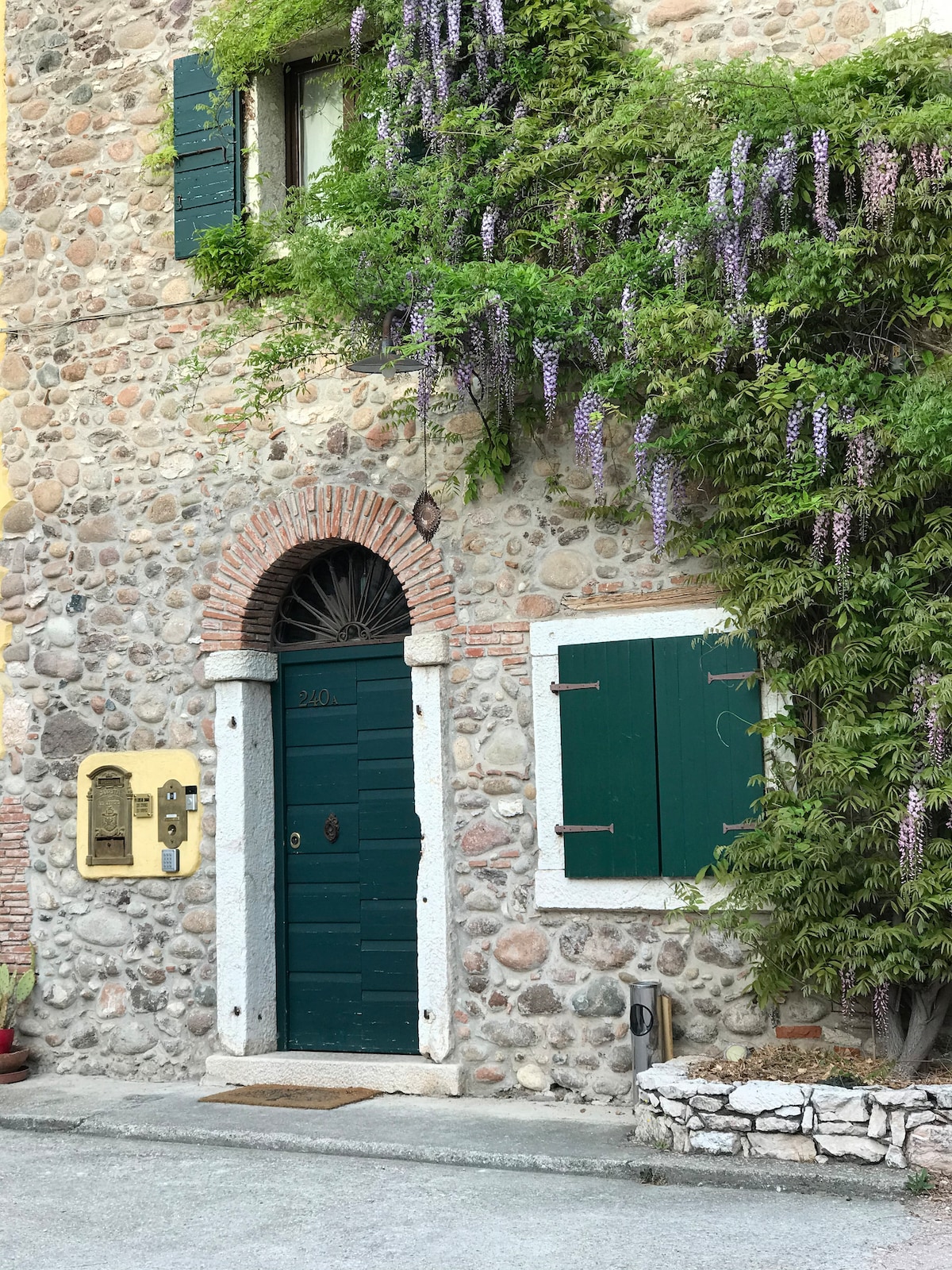 A rustic stone facade presents a welcoming entrance, featuring a central wooden door framed by greenery and vibrant purple wisteria. Flanked by green shutters, an intercom is mounted beside the door, indicating the entrance to the inviting property.