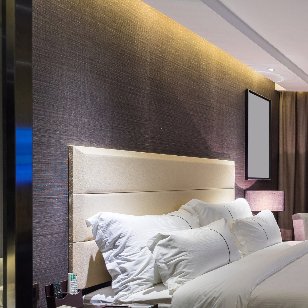 A comfortable bedroom setting features a light-colored headboard against a textured dark wall. Crisp white pillows are arranged on a spacious bed, and a bedside table is visible with a remote control. Soft lighting highlights the warm tones of the room.