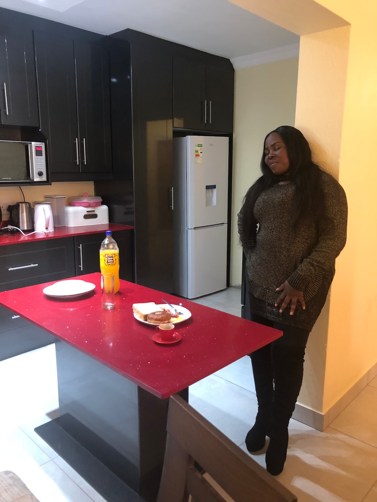 A modern kitchen features sleek black cabinetry and a vibrant red countertop. On the island, plates hold food items, while a bottle of soda adds a pop of color. Stainless steel appliances are visible, with a microwave and refrigerator contributing to a functional design.