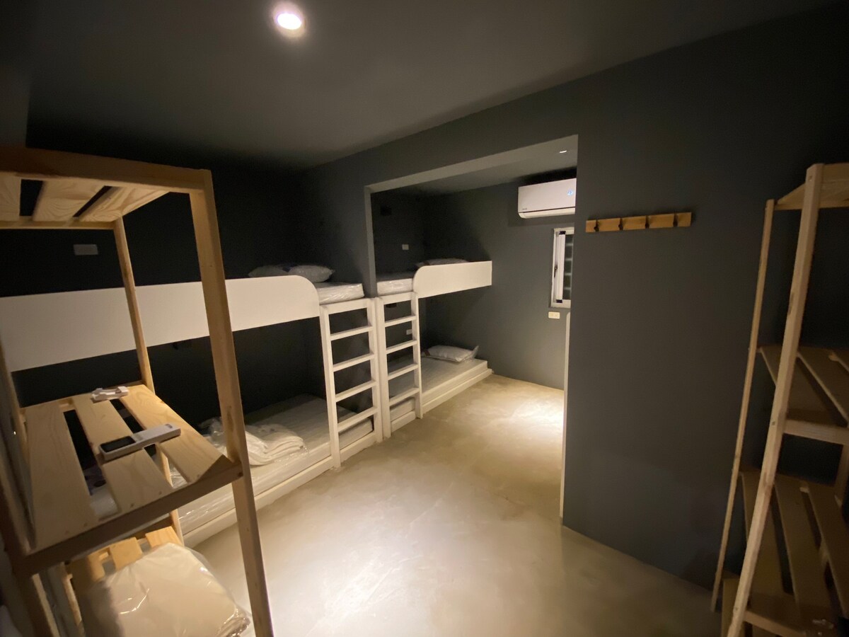 A shared sleeping area contains two sets of bunk beds, with each bed arranged against a dark wall. Additional wooden shelves are positioned along one side, providing storage space. Soft lighting illuminates the room, enhancing the overall simplicity and functionality.