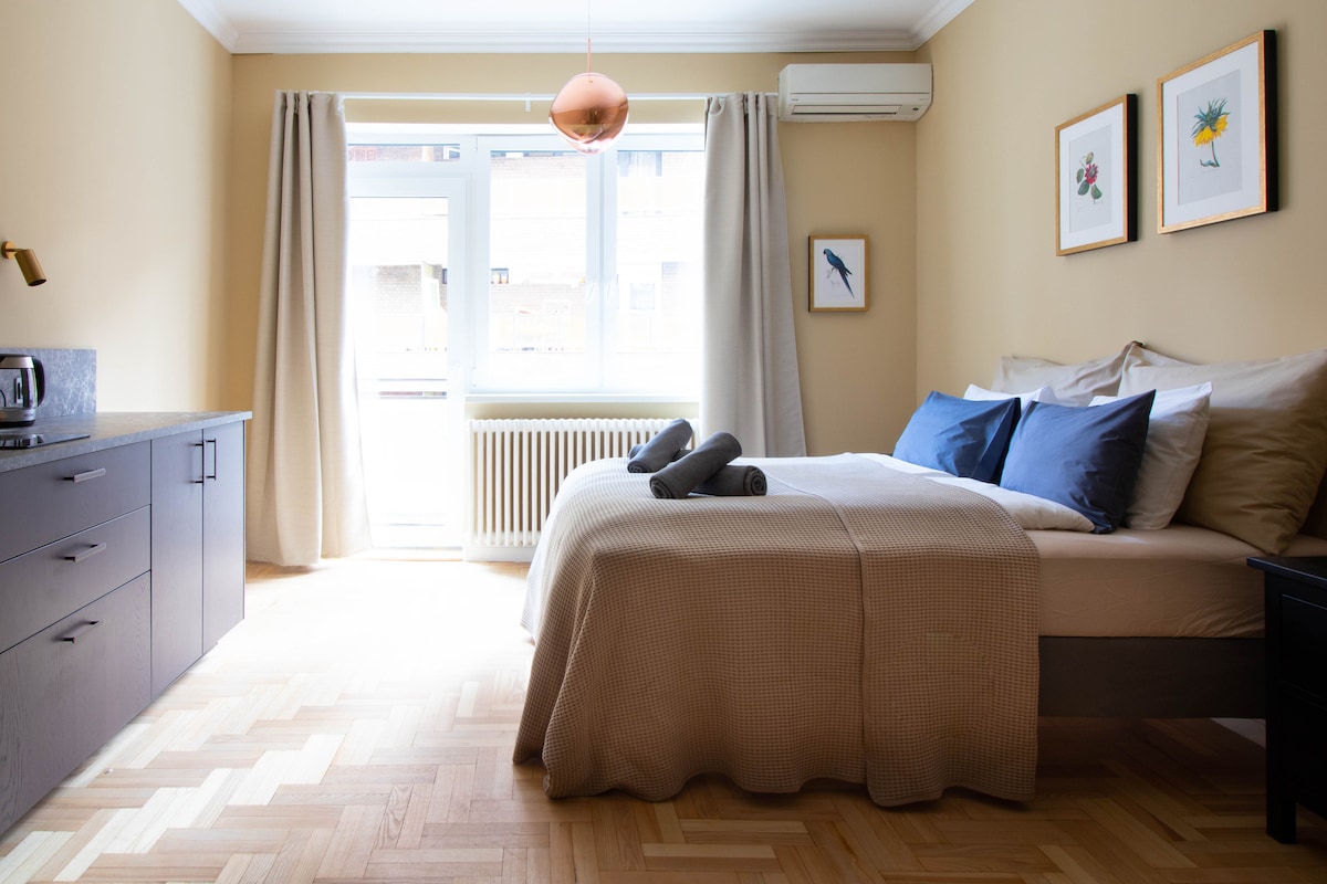 A spacious bedroom features a neatly made bed with blue pillows and rolled towels placed on a light-colored blanket. Natural light floods in through large windows, complemented by soft beige walls and decorative artwork. A compact kitchenette is visible in the corner.