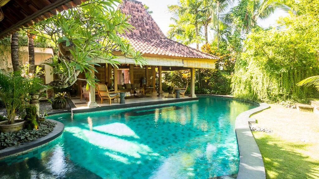 The image features a tranquil lagoon pool, surrounded by lush tropical vegetation. A traditional Indonesian Joglo structure stands nearby, its roof topped with terracotta tiles. Sunlight filters through the greenery, casting dappled light on the water's surface.