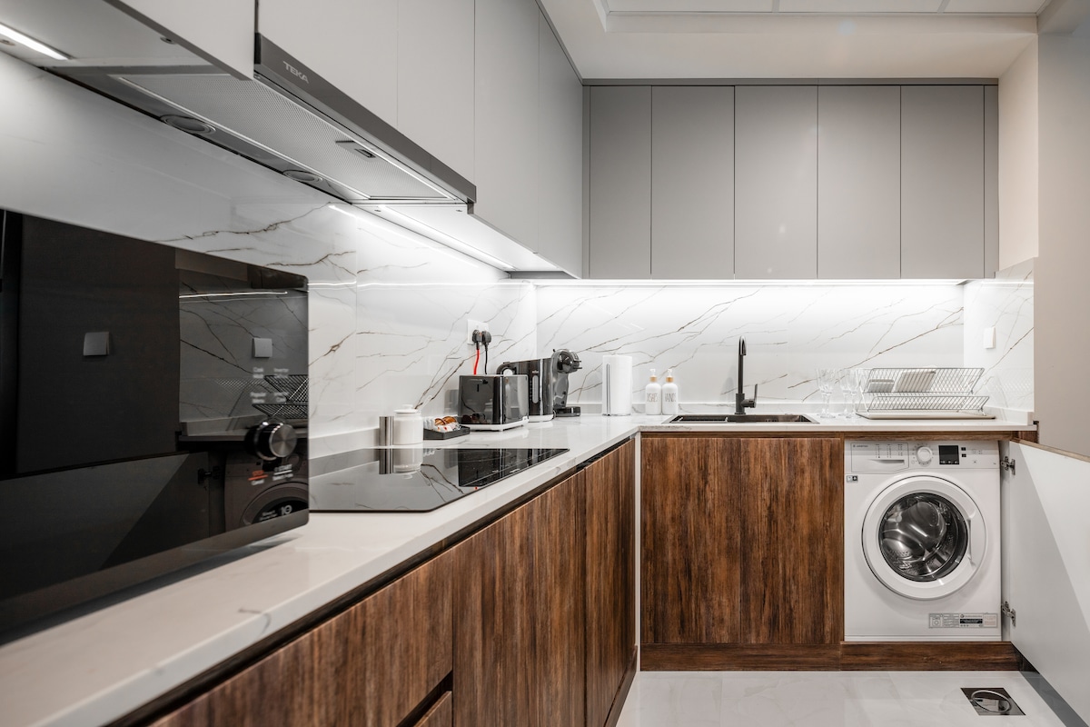A modern kitchen is displayed with a mix of sleek grey cabinetry and warm wooden accents. Appliances include a built-in microwave, induction stove, and a washing machine. Under-cabinet lighting enhances the counter space, which is equipped with a sink and dish drying rack.