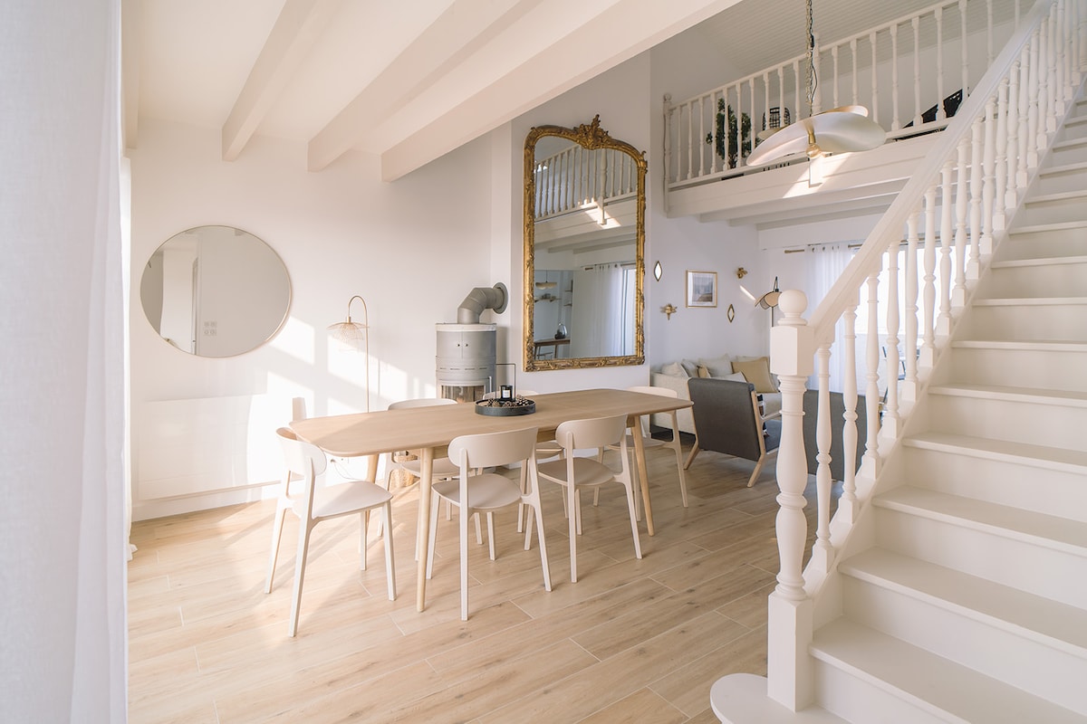 A bright dining area is complemented by a long wooden table surrounded by six chairs. A large mirror hangs on the wall, reflecting natural light from the windows. A staircase leads to the upper level, and a soft seating area is visible in the background.