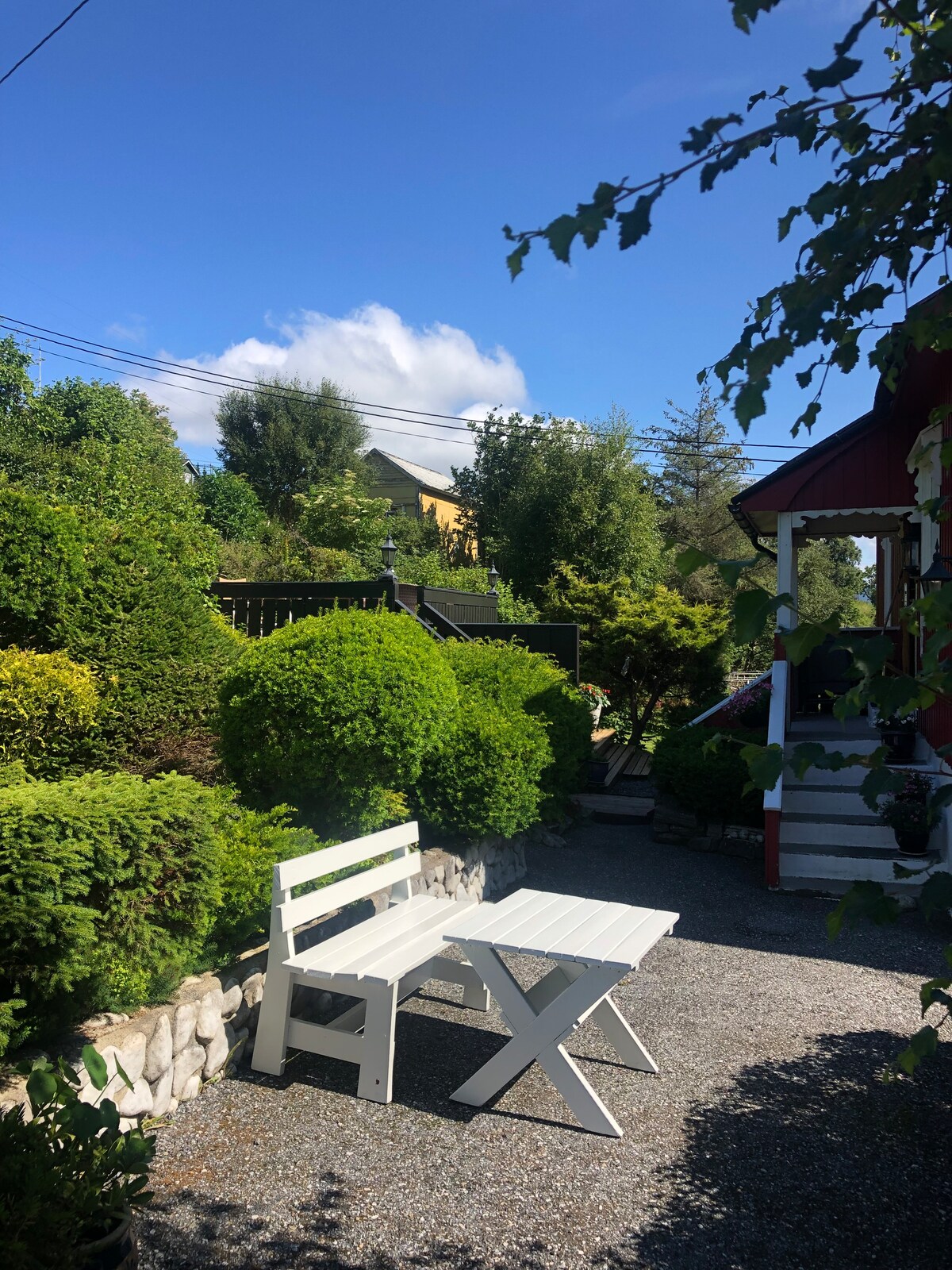 A well-maintained garden area is depicted, featuring neatly trimmed hedges and lush greenery. A white wooden bench and table are positioned on a gravel pathway, inviting relaxation. Bright blue skies and fluffy clouds overhead enhance the natural setting.