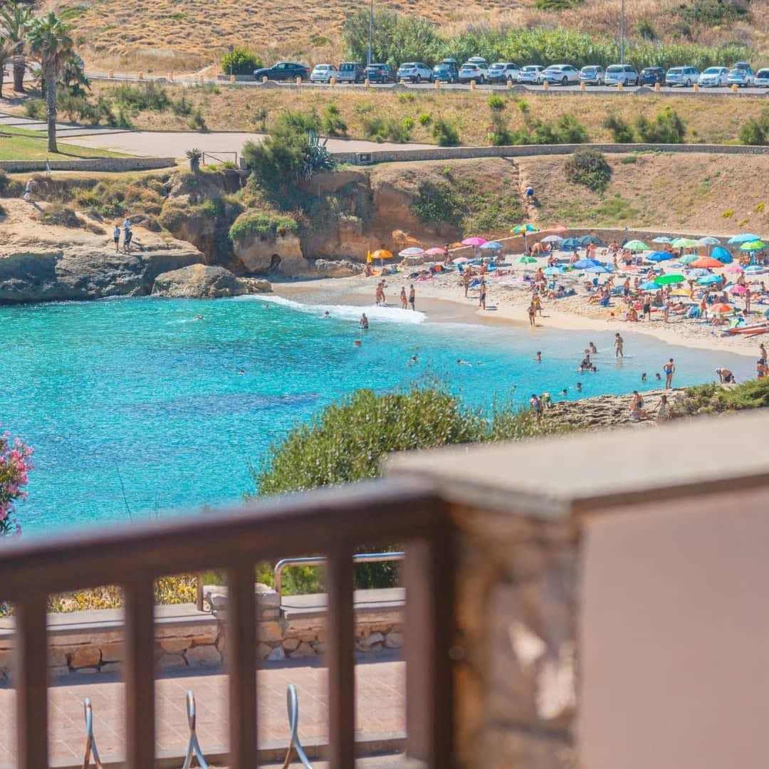 A view from a balcony captures the vibrant beach scene, where colorful umbrellas dot the sandy shore and people enjoy the turquoise waters. Green shrubs frame the foreground, and a roadway with parked cars is visible in the background, emphasizing the proximity to the beach.