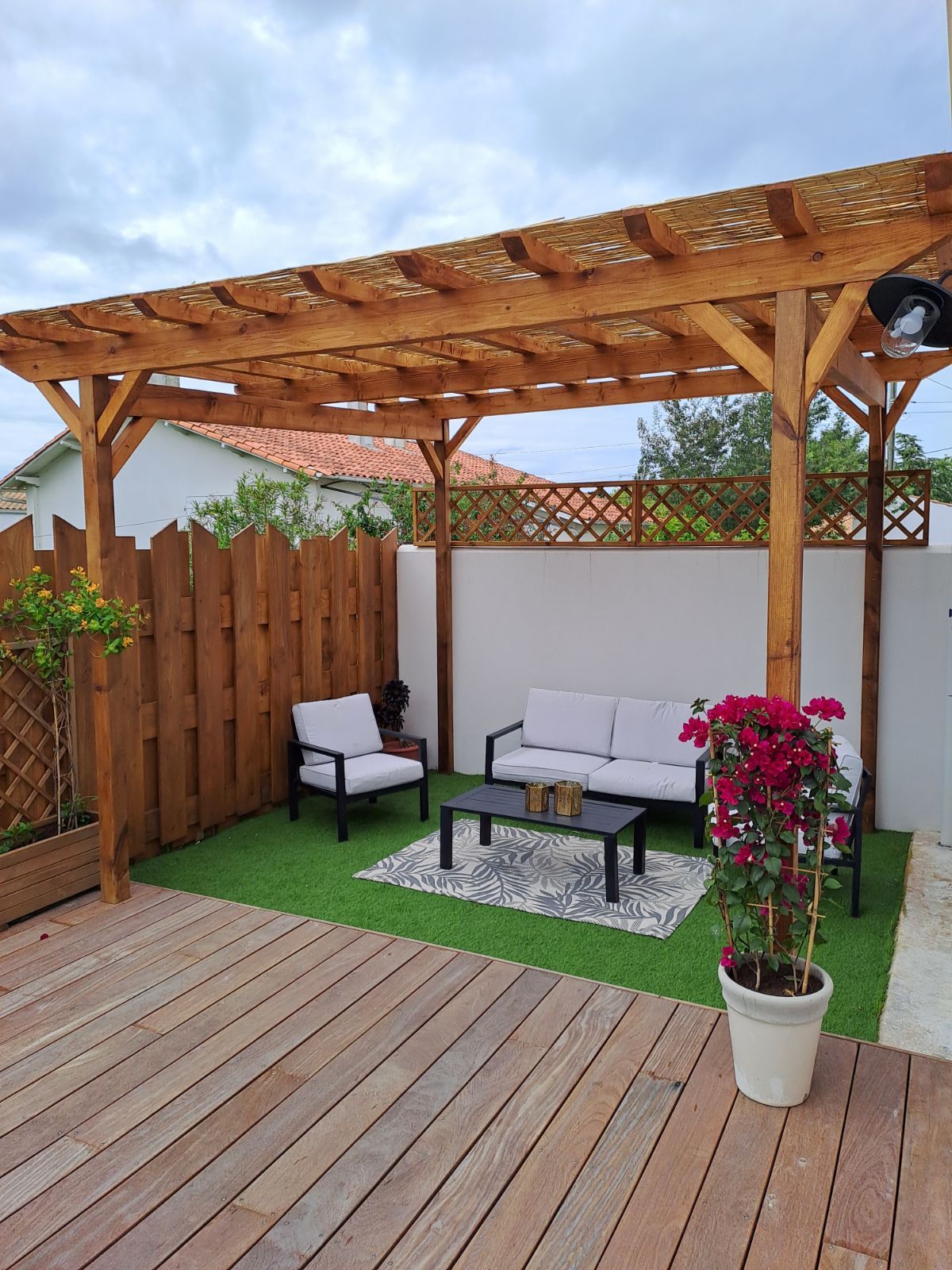 An outdoor terrace is presented, featuring a wooden pergola overhead. Two white chairs and a matching sofa are arranged around a dark coffee table, all set on a patterned rug. Potted flowers add a touch of color, while wooden fencing provides privacy.