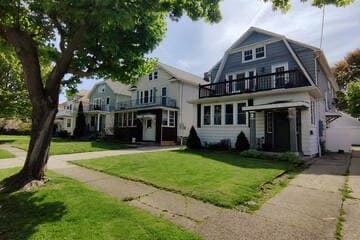 An inviting view of a tree-lined street featuring two white and gray upper units of a two-family home. Lush green lawns surround the properties, and balconies are visible on the upper levels, enhancing the welcoming neighborhood ambiance.