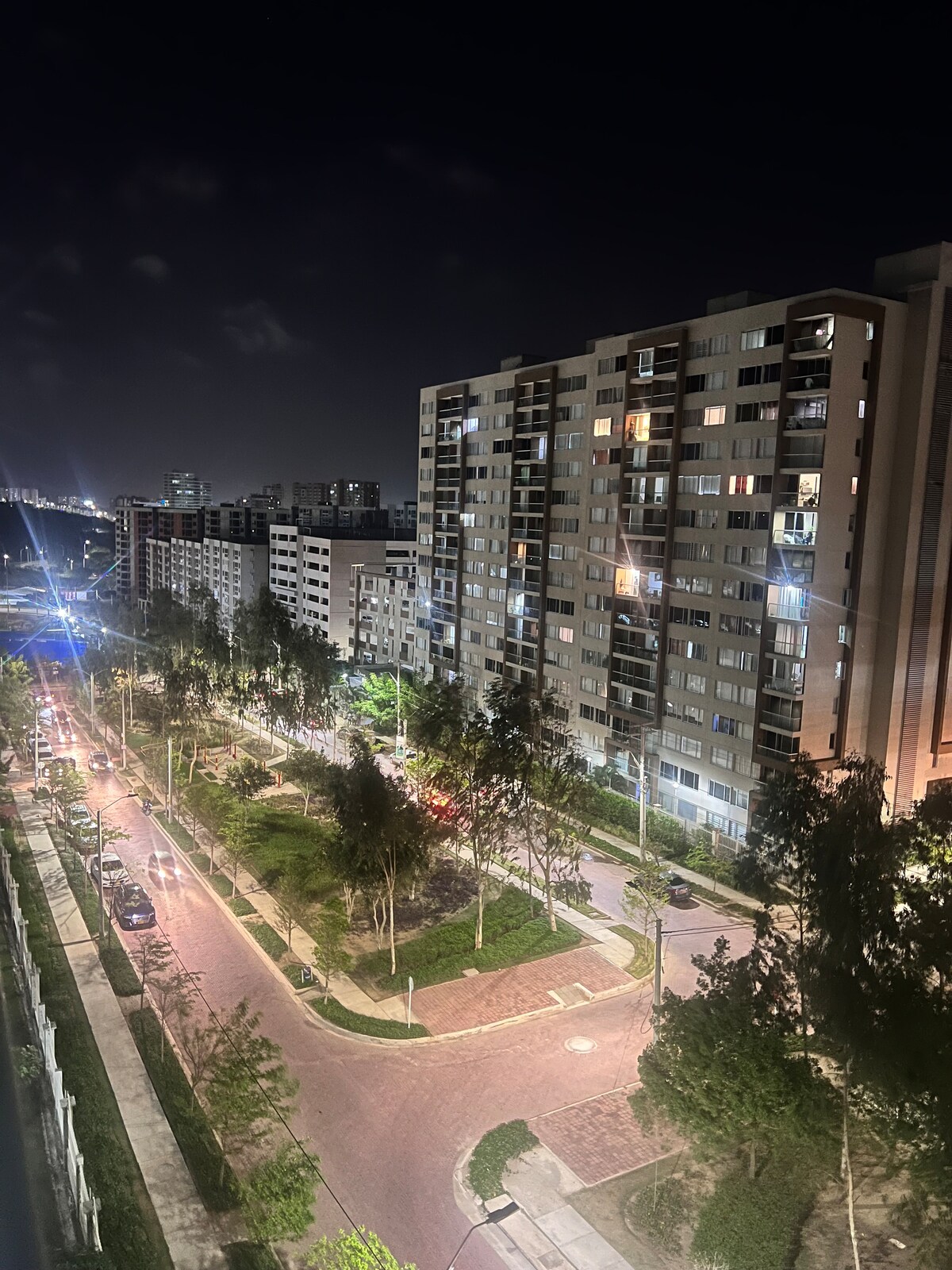 A cityscape is depicted at night, showcasing a well-lit street lined with trees and several apartment buildings. The vibrant lights from the surrounding structures create a lively atmosphere, highlighting the urban environment. The road curves gently, leading towards a distant view of the city skyline.