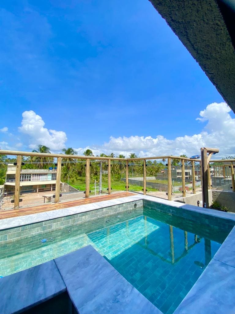 A private rooftop pool reflects the blue sky, surrounded by a wooden railing. Lush greenery is visible in the distance, enhancing the outdoor view and providing a serene atmosphere.