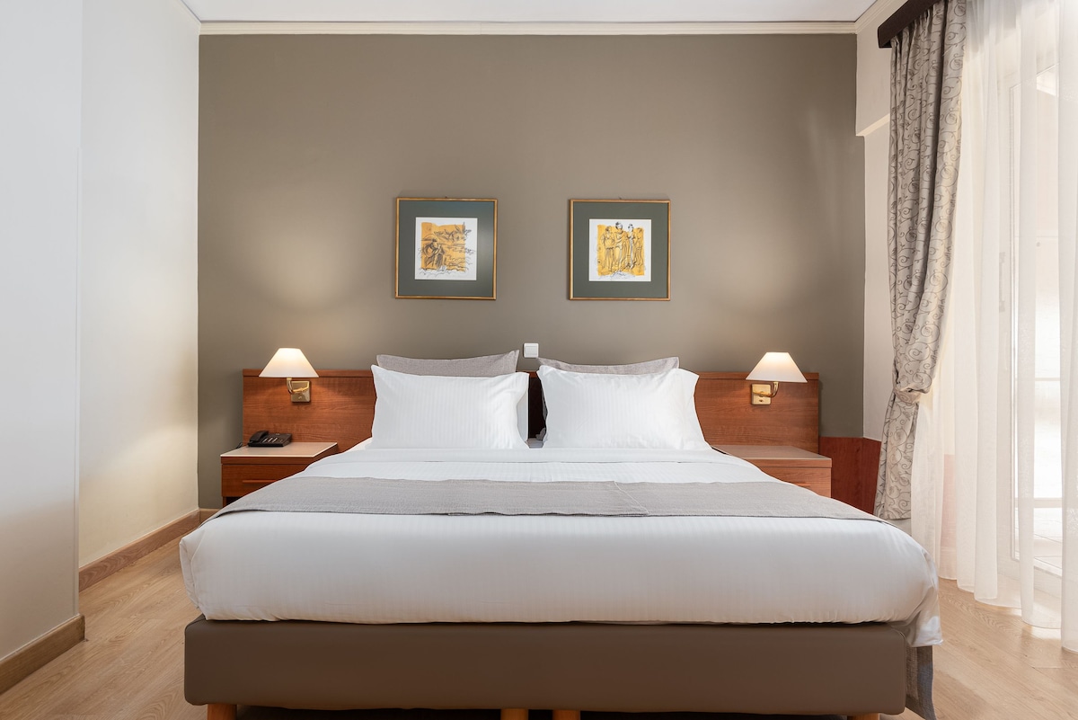 A spacious room features a large bed topped with crisp white linens, complemented by elegant wooden furniture. Soft lighting is provided by bedside lamps, while tasteful artwork adorns the walls. Natural light enters through sheer curtains, creating a peaceful ambiance.