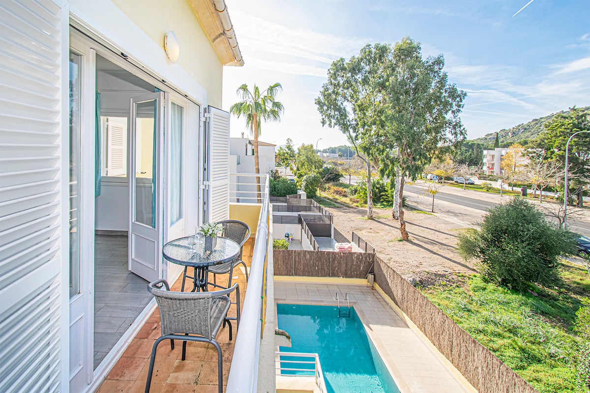 A private balcony showcases a small outdoor table with two chairs, overlooking a pool surrounded by sunlit greenery. The view includes a quiet street and distant hills, with sliding glass doors leading into the interior space.
