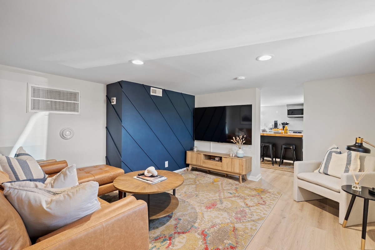 The living area features a cozy leather sectional sofa arranged around a circular coffee table. A colorful area rug lies underneath, contrasting with the light wood flooring. A mounted flat-screen TV is positioned against a stylish blue accent wall, creating a modern and inviting space.