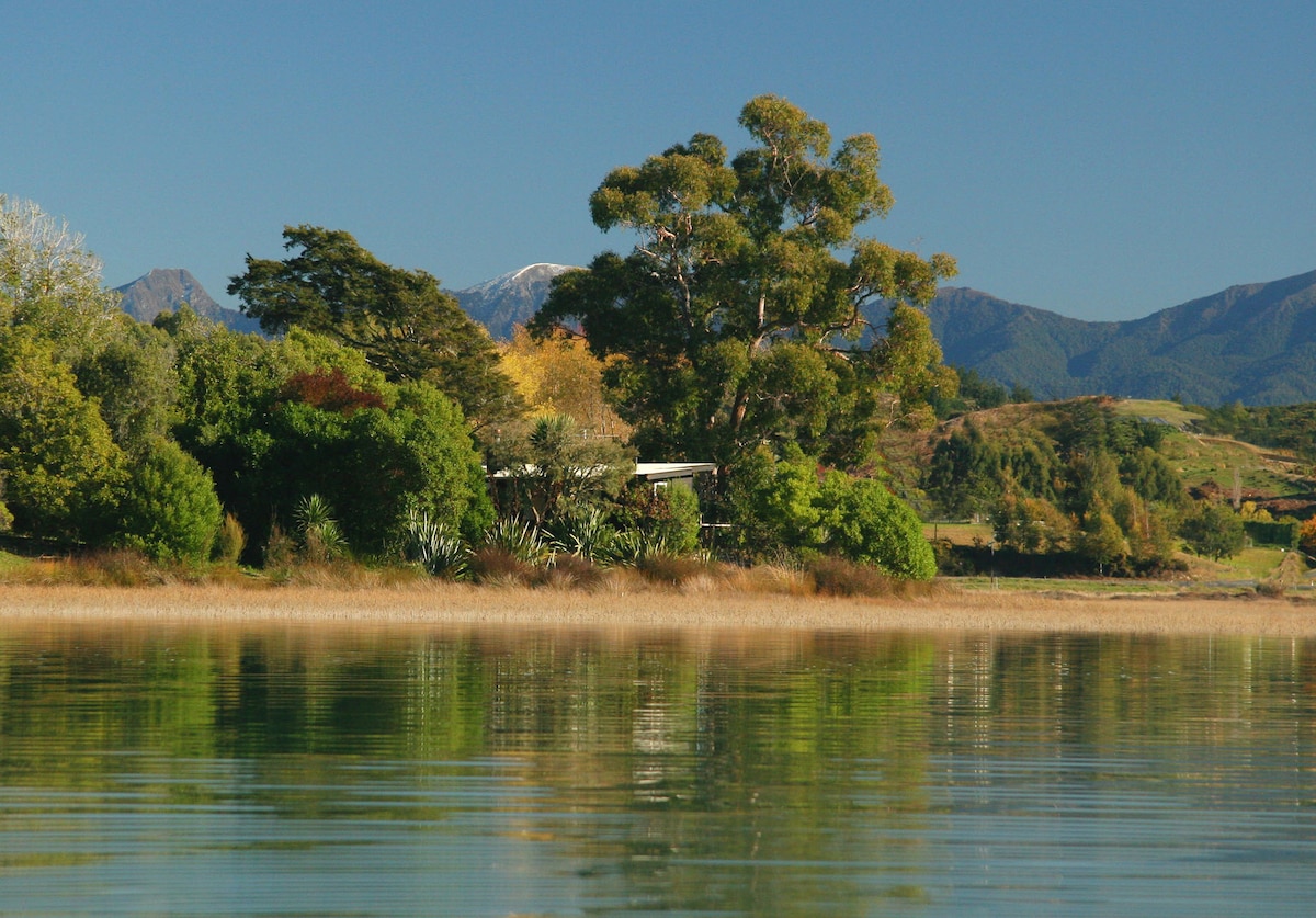 The image captures a serene landscape with a reflection of lush trees along the water's edge. The backdrop features distant mountains, partially covered with snow, under a clear blue sky. The tranquility of the scene is enhanced by the calm surface of the water.