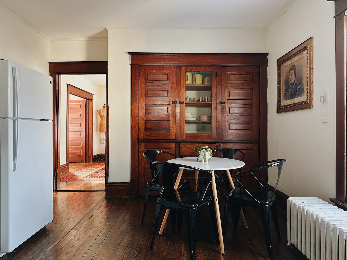 A well-lit kitchen area features a small round table surrounded by black chairs, complemented by a bouquet at its center. The wooden cabinetry showcases glass-fronted sections, creating a warm contrast with the white refrigerator. Doors leading to other spaces are visible in the background.