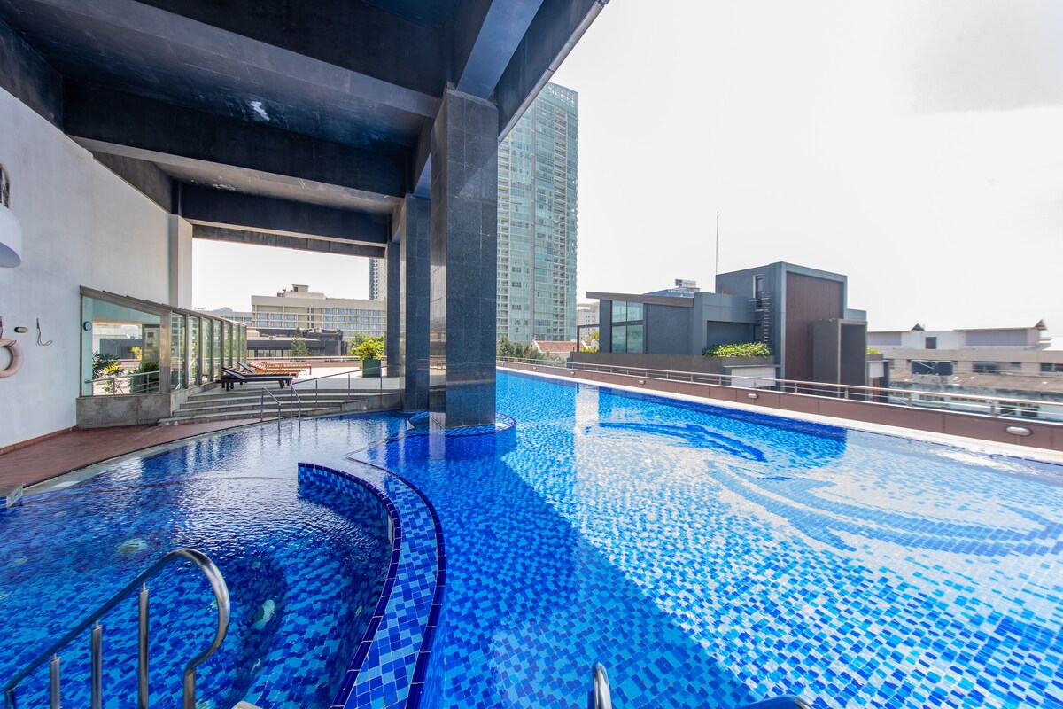 A rooftop swimming pool is presented, featuring a vibrant blue mosaic tiled surface. Sunlight reflects off the water, creating a shimmering effect. Surrounding areas include comfortable seating and views of the surrounding skyline, enhancing a sense of openness and relaxation.