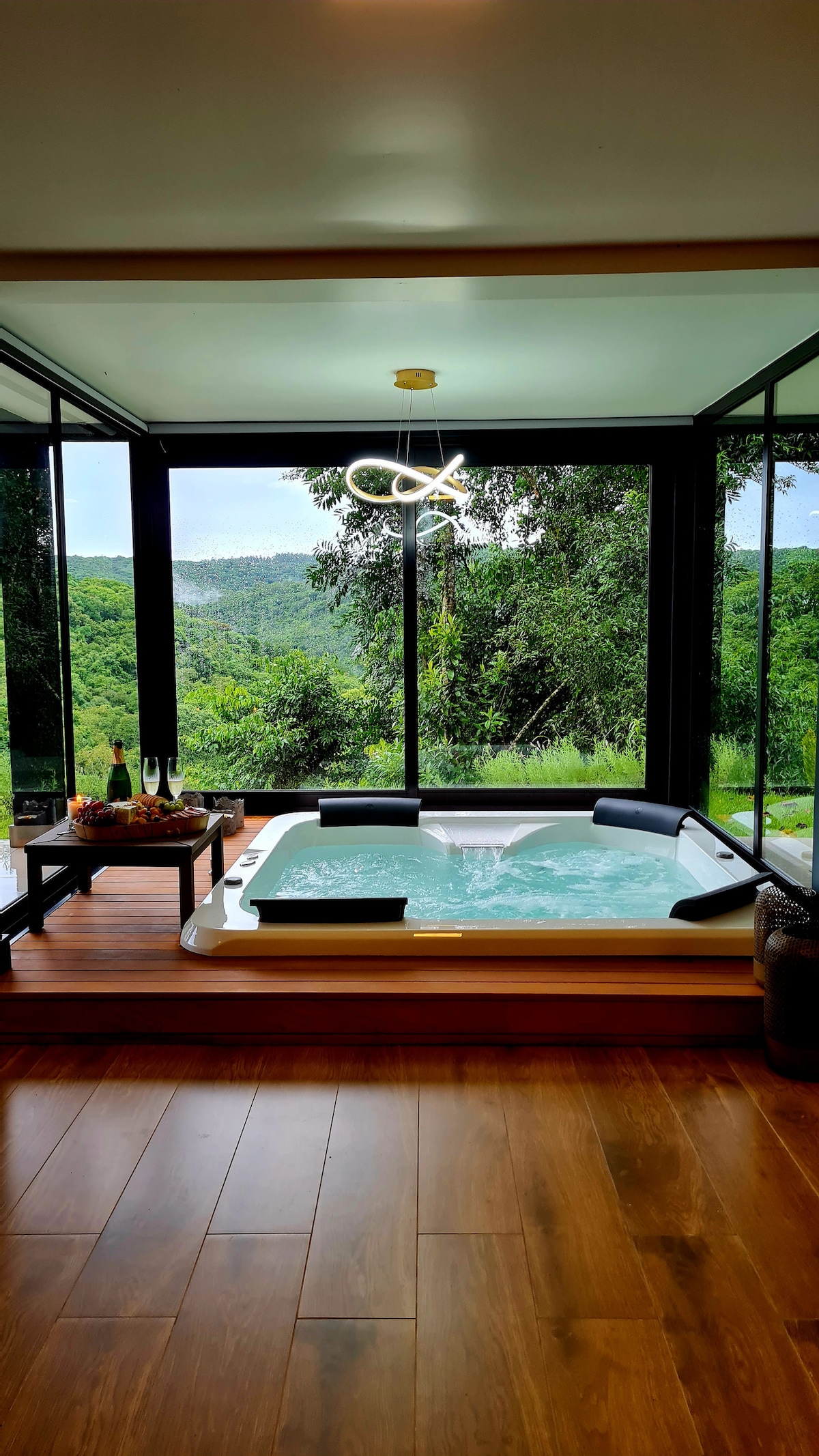 A spacious jacuzzi is situated within a glass-enclosed area, offering views of the surrounding greenery. Natural light illuminates the room, which features wooden flooring and a stylish modern décor. The ambiance is complemented by a unique overhead light fixture.
