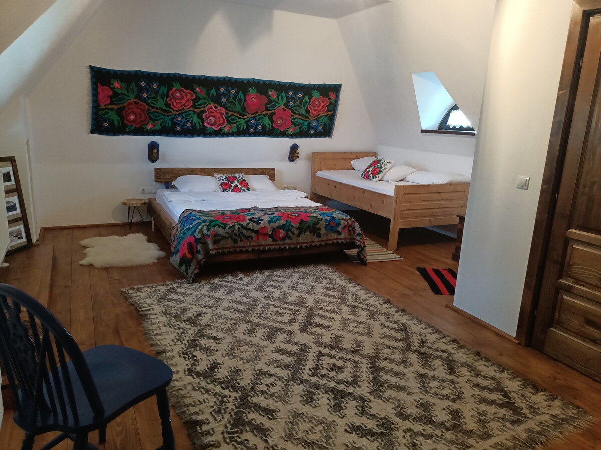 A spacious room features two wooden beds, one larger and one smaller, adorned with decorative blankets. A traditional textile artwork hangs above, providing a colorful accent. Soft area rugs are placed on the wooden floor, enhancing the room's rustic charm. Natural light enters through a window.