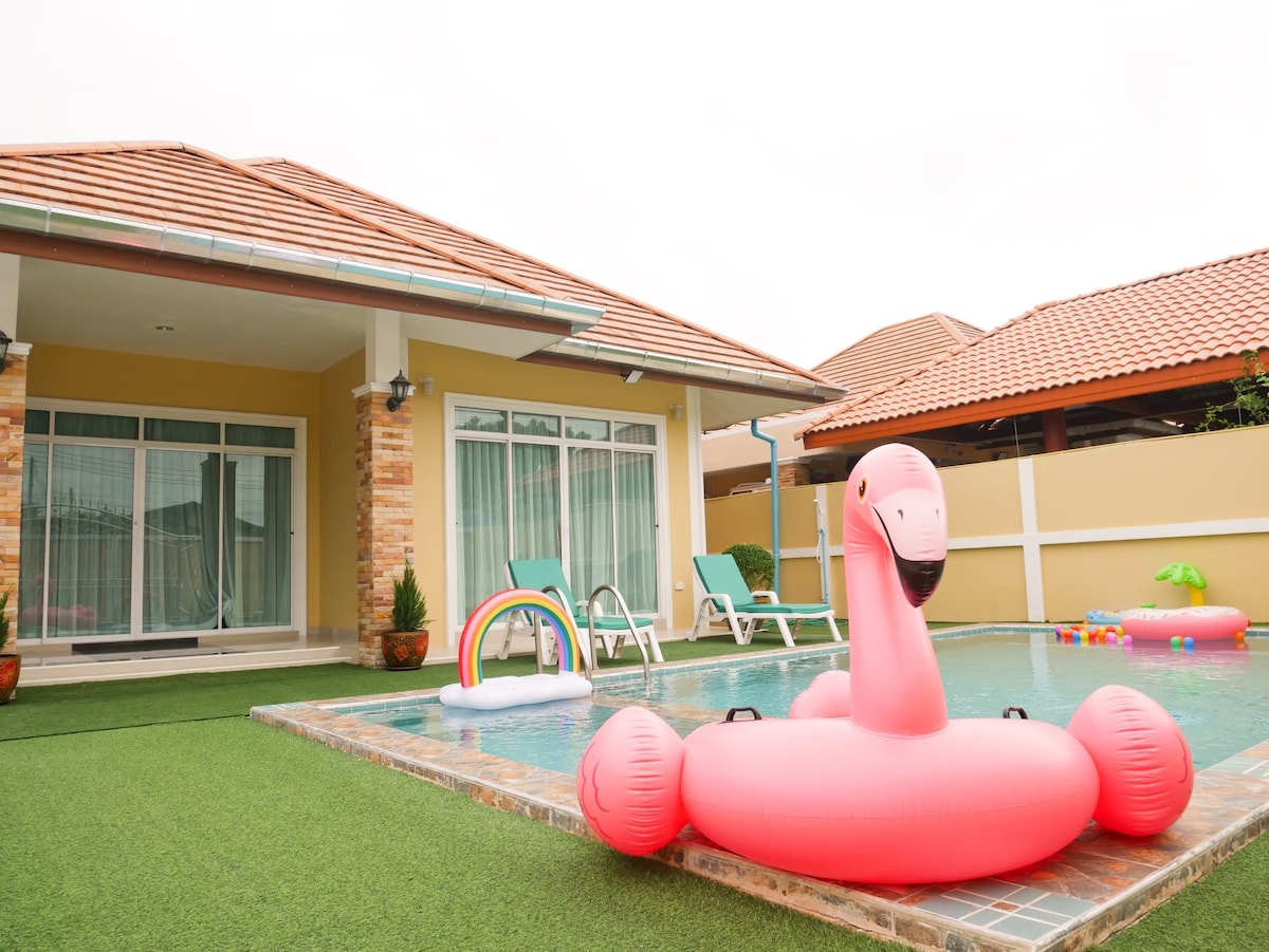 A vibrant swimming pool area features a large pink inflatable flamingo in the water. Nearby, lounge chairs with green fabric are placed on manicured grass. The villa’s exterior is seen in the background, showcasing a warm color palette and an inviting atmosphere.