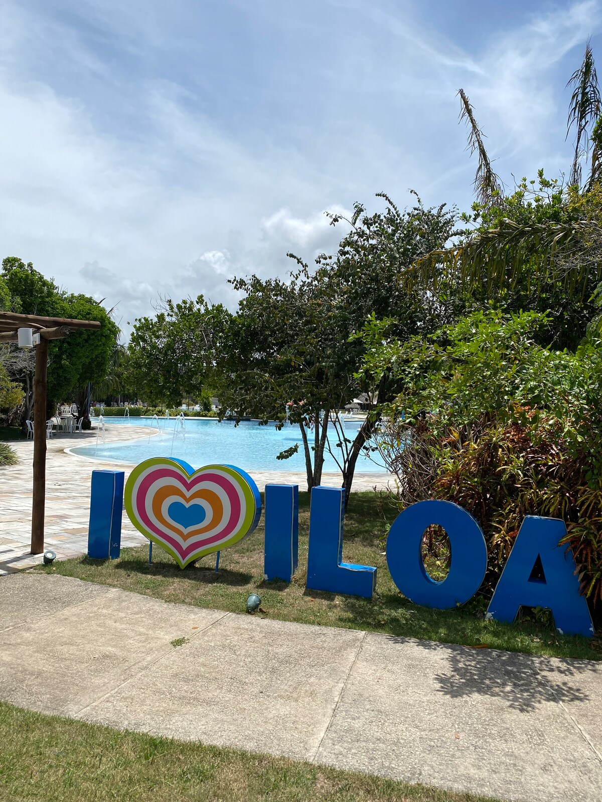 A colorful 'I Love Iloilo' sign is prominently displayed in the foreground, surrounded by lush greenery. In the background, a large outdoor pool is visible, offering a refreshing retreat under a bright sky.