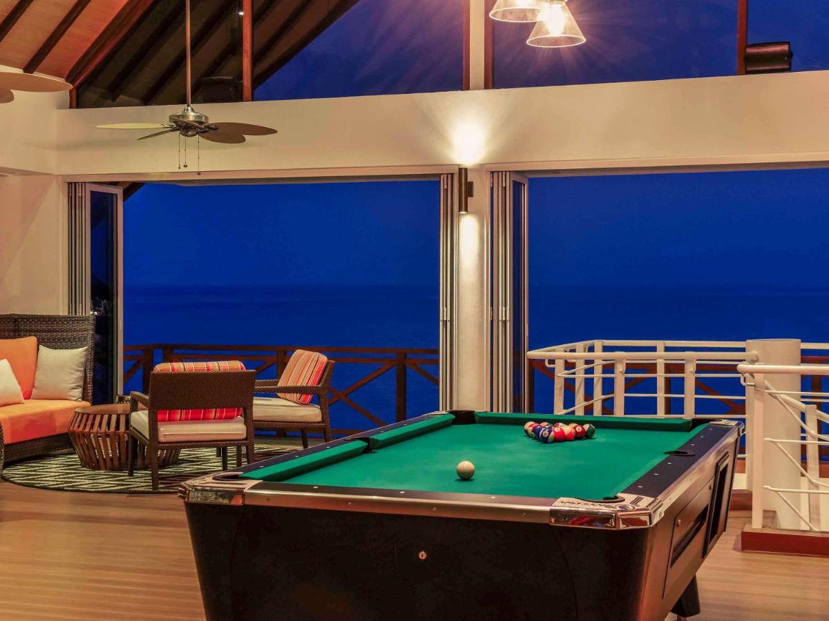 A pool table is positioned centrally in the room, surrounded by comfortable seating. Large glass windows offer unobstructed views of the water, creating a bright ambiance as evening falls. The ceiling is adorned with a ceiling fan, and the decor features a blend of modern furnishings and warm colors.