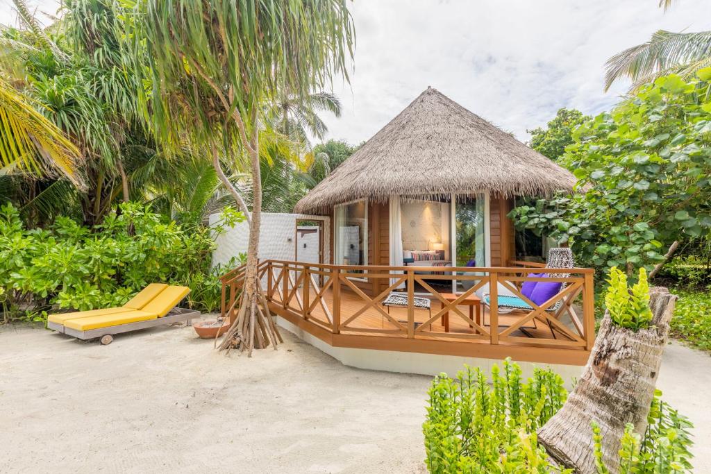 A beach villa featuring a thatched roof is set amidst lush greenery, with a spacious wooden deck visible in the foreground. Brightly colored lounge chairs are arranged on the sandy area, providing a place to relax in the tropical surroundings.