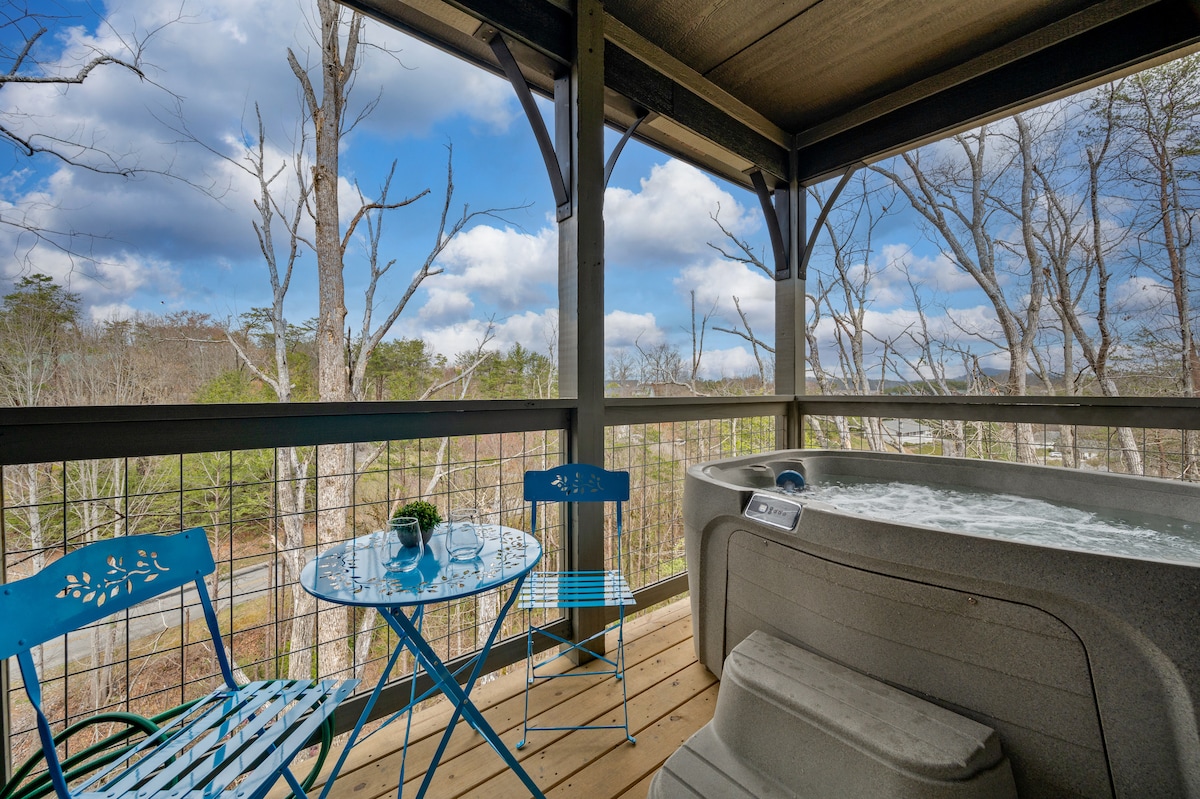 A private deck features a hot tub and a bistro dining table with two blue chairs. The surrounding trees offer a natural view, while the open sky above showcases a blend of clouds. The space is designed for relaxation and enjoyment of the outdoors.