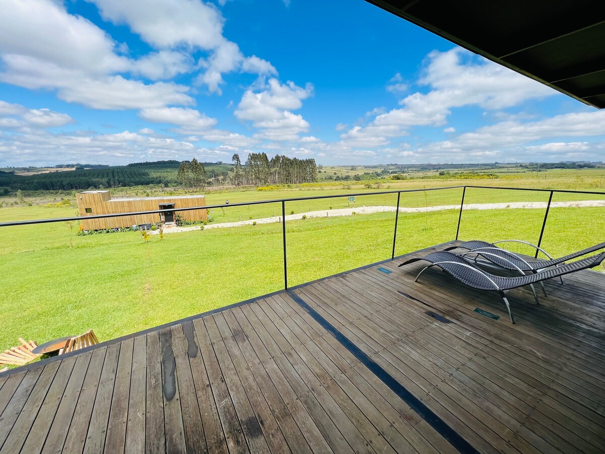 A wooden deck features a serene view of expansive grassy fields and rolling hills under a partly cloudy sky. A pair of modern lounge chairs is positioned for relaxation, offering a peaceful outdoor space surrounded by nature.