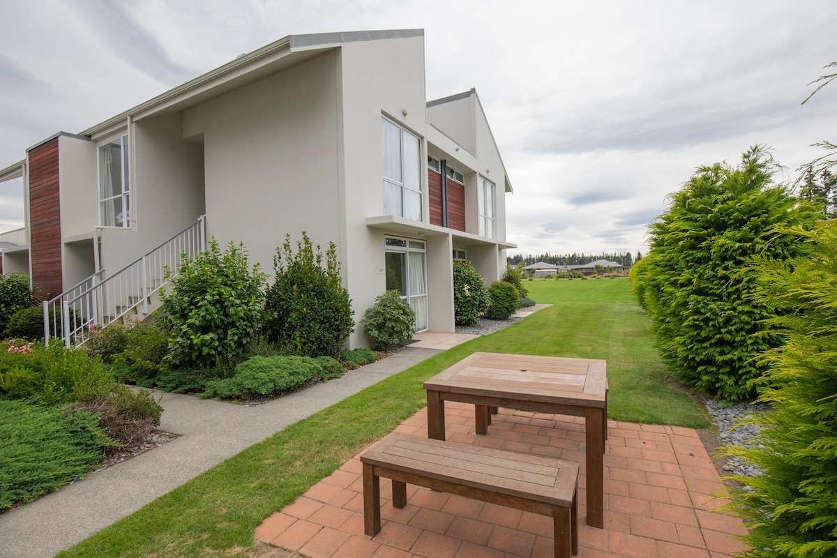 The exterior of an apartment complex is viewed, featuring a garden area with well-maintained greenery. A wooden picnic table is positioned on a paved patio, providing an outdoor relaxation space. The building has two levels, with large windows allowing natural light to enter the units.
