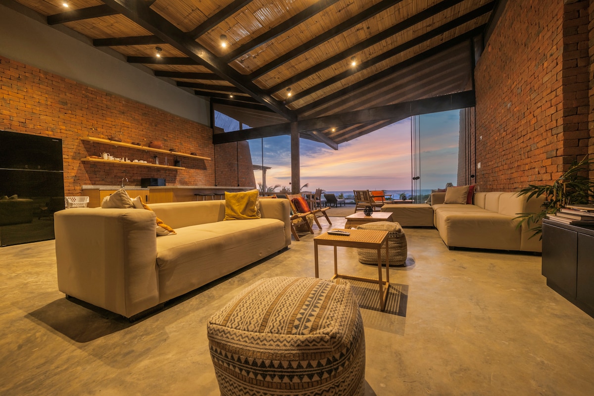 A spacious living area features large glass doors that open towards a sunset view. Soft, neutral-colored sofas are arranged around a central coffee table. Exposed brick walls add texture, while the ceiling showcases wooden beams, contributing to an open and airy atmosphere.