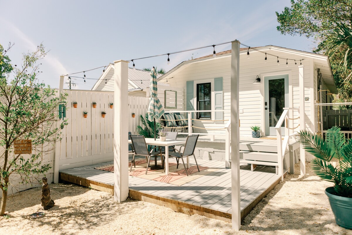 An outdoor deck area is featured, equipped with a table and four chairs. Surrounding greenery includes plants and a decorative statue. A white fence offers privacy, with string lights adding ambiance. The cottage exterior reflects a clean, modern design, complemented by coastal elements.