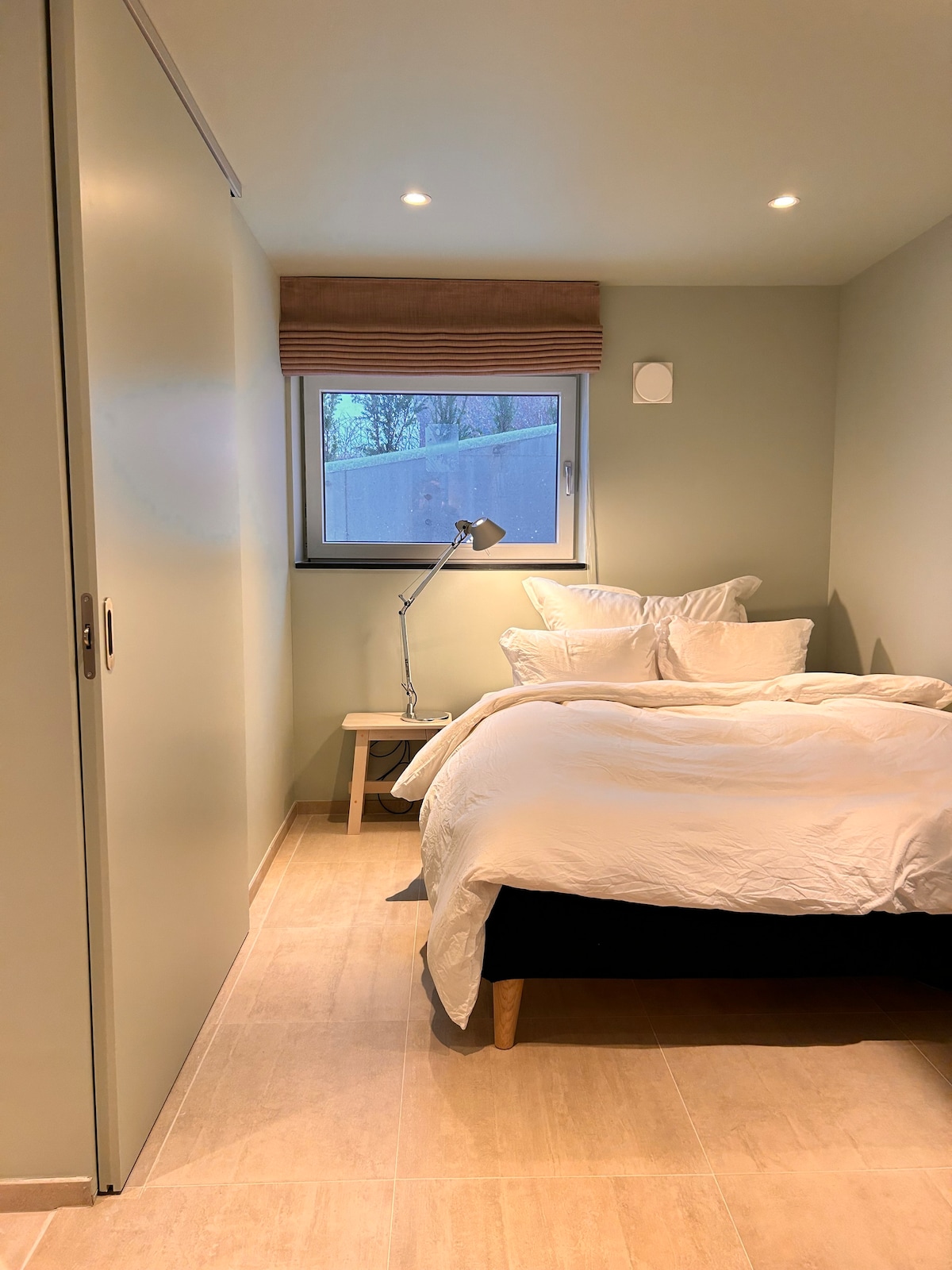 A cozy bedroom is shown, featuring a spacious bed with white linens and several pillows. Natural light enters through a window adorned with a simple blind. A small bedside table is present, alongside a modern lamp for added illumination. A sliding door leads to additional space.