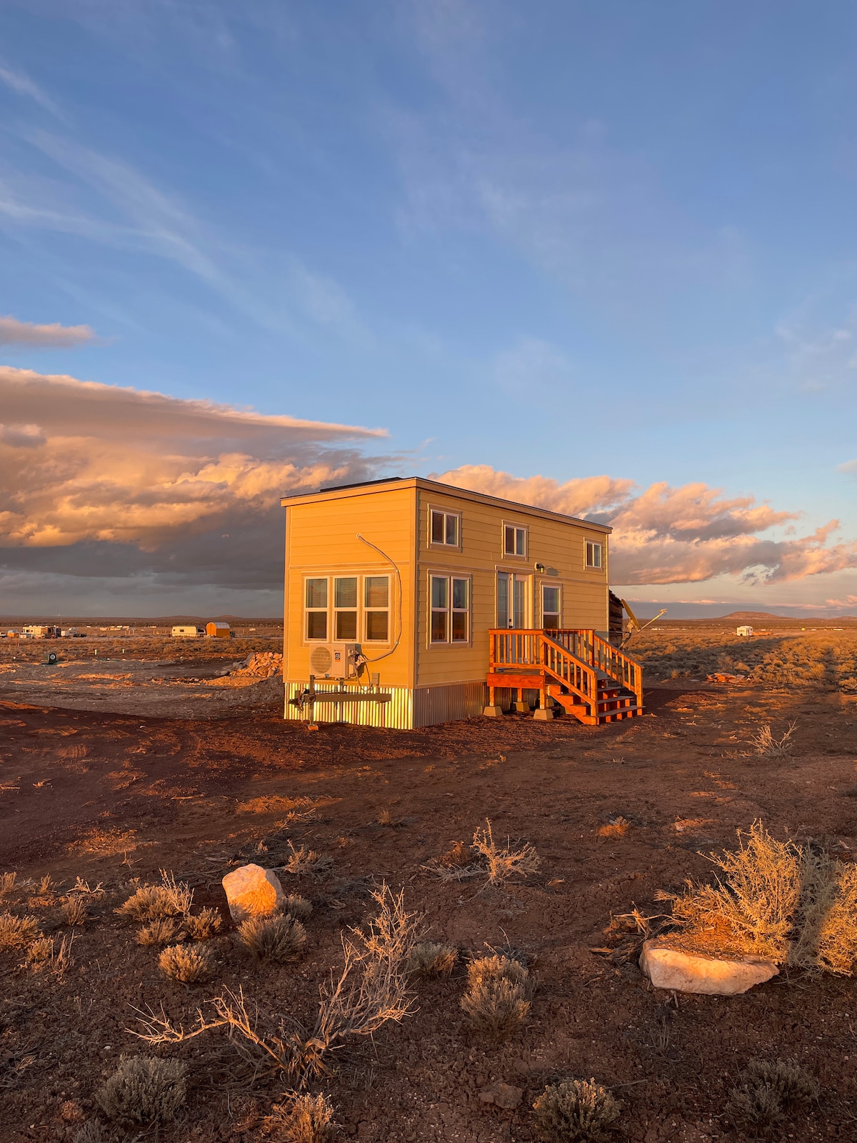 A cozy yellow tiny home is showcased in a vast landscape under a colorful sky, illuminated by the warm glow of sunset. The exterior features multiple windows and a wooden staircase leading to the entrance, surrounded by arid terrain and scattered rocks.