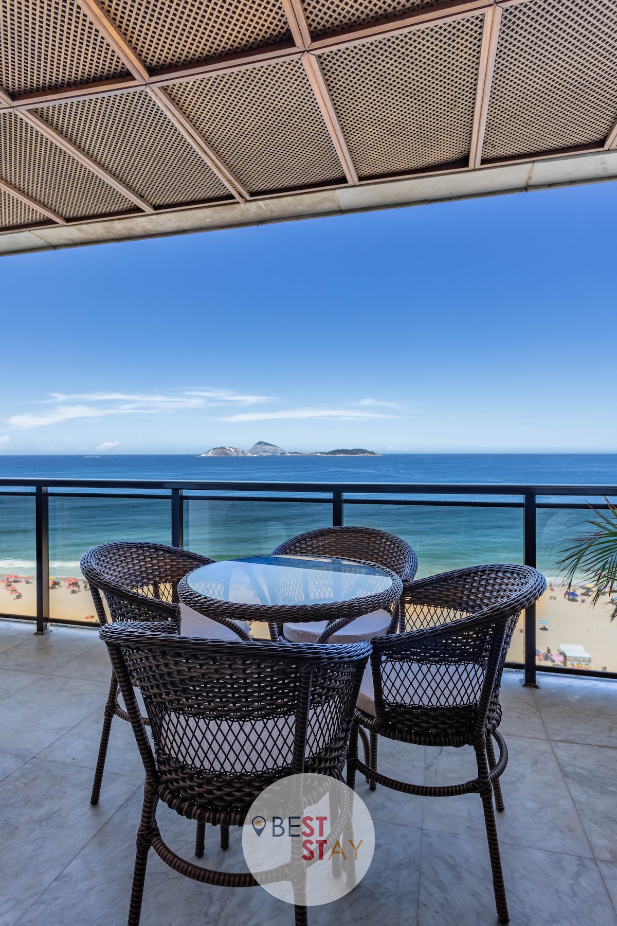 A balcony features a table surrounded by four woven chairs, offering stunning views of the ocean and beach below. The horizon stretches into a clear blue sky, complemented by distant landforms that enhance the scenic backdrop.