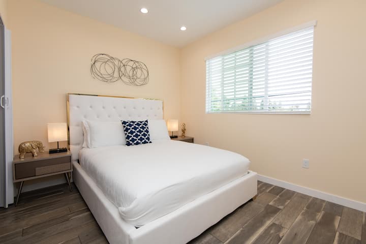 Светла и уютна спалня с бяло легло с декоративни възглавници и меко осветление.