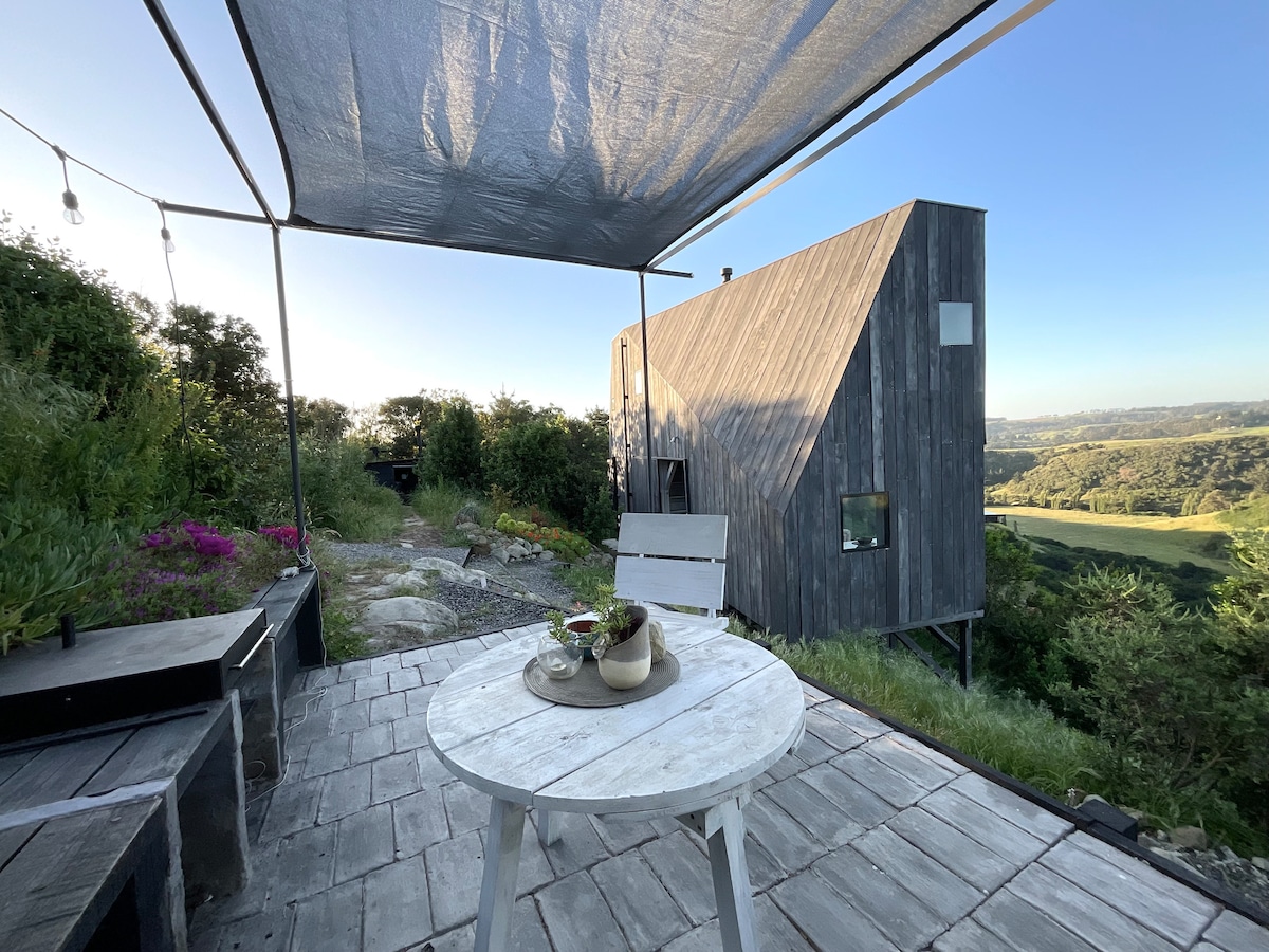 An outdoor patio area features a round white table with light seating beneath a shaded canopy. Lush green landscape and vibrant flowers are visible in the background, with a unique wooden structure rising in the distance. Soft evening light enhances the tranquil setting.