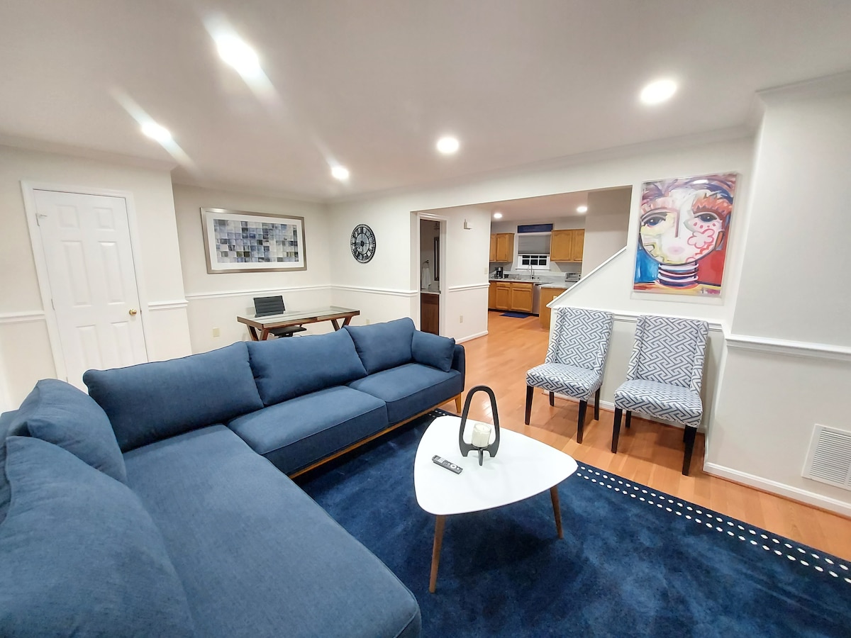 A spacious living area is presented, featuring a large blue sectional sofa and a light coffee table atop a navy area rug. Additional seating includes two patterned chairs. Artwork decorates the walls, and a desk is positioned nearby, along with a view of the kitchen area.