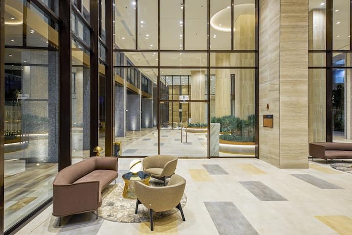 A spacious lobby is designed with tall glass walls, allowing natural light to fill the area. Comfortable seating is arranged in a conversational layout, complemented by modern decor and potted greenery. The floor features a combination of smooth tiles, enhancing the contemporary aesthetic.