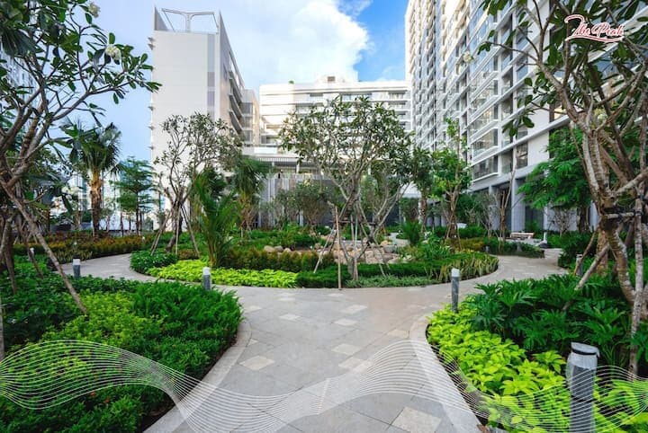 A landscaped garden area is surrounded by modern condominium buildings. Pathways are lined with lush greenery and trees, creating a serene environment. Well-maintained flower beds add vibrant colors to the scene, while soft lighting fixtures are placed along the paths.
