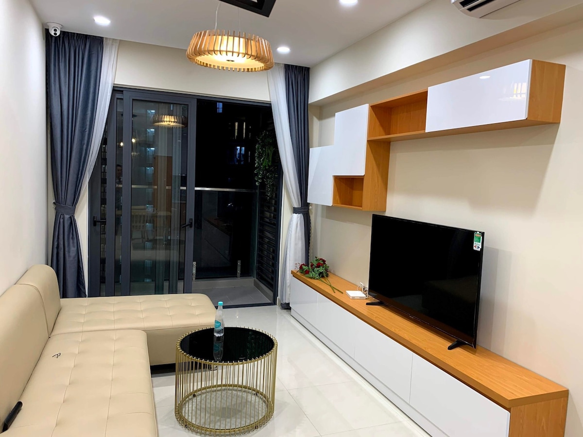 A modern living area is presented with a light-colored sectional sofa and a round glass coffee table. A minimalist entertainment unit holds a flat-screen TV, while large glass doors provide access to a balcony. Soft drapes frame the windows, adding a touch of elegance.