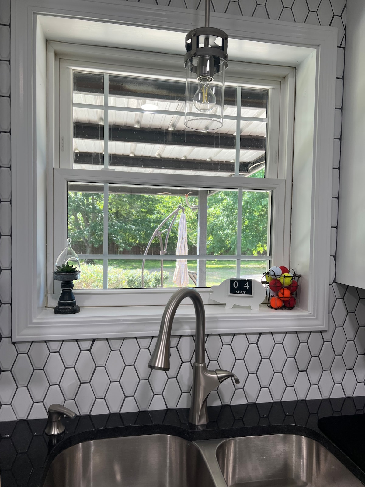 A modern kitchen window offers a view of the backyard, framed by white trim. Natural light enhances the light hexagon tile backsplash. A stylish faucet and stainless steel sink are visible, with a plant and colorful items placed on the windowsill, adding a touch of homeliness.