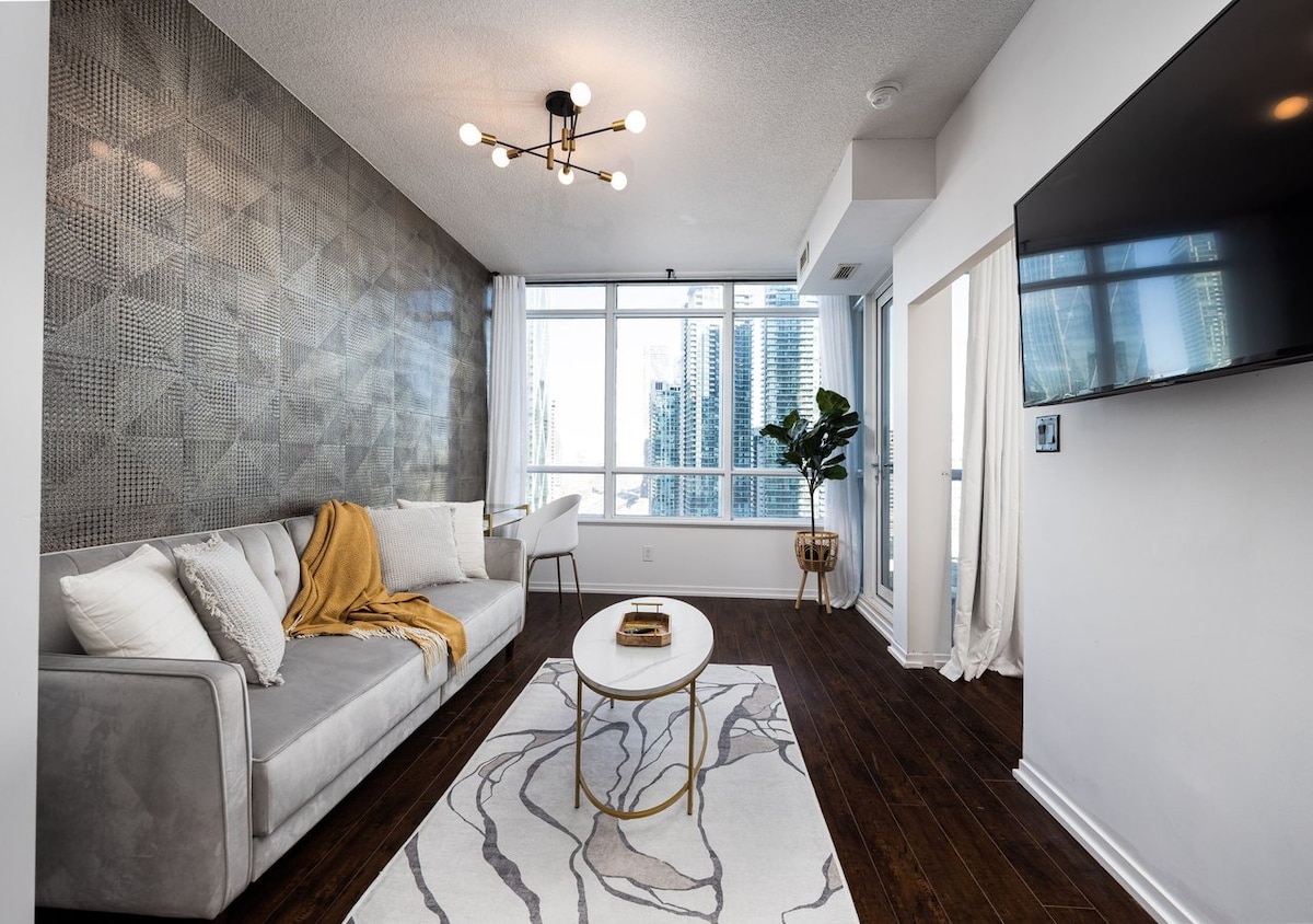 A stylish living area features a sleek, grey sofa adorned with a warm throw. A round coffee table rests on a patterned rug, while large windows provide an abundance of natural light and views of the city skyline. A modern light fixture hangs above, enhancing the contemporary design.