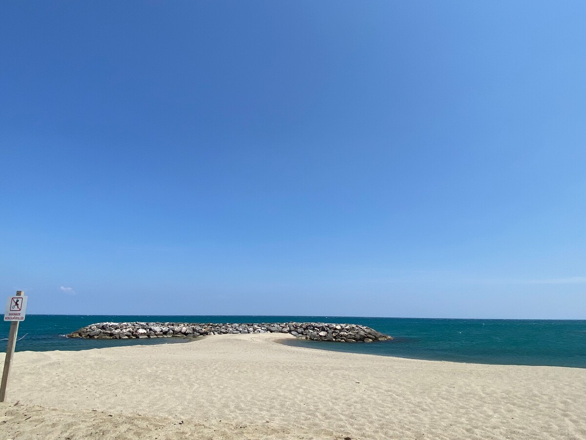 A sandy beach stretches toward a peaceful body of water, with gentle waves lapping against a rocky jetty. The sky is clear and blue, creating a serene coastal atmosphere. A sign stands at the edge of the beach, indicating swimming rules.