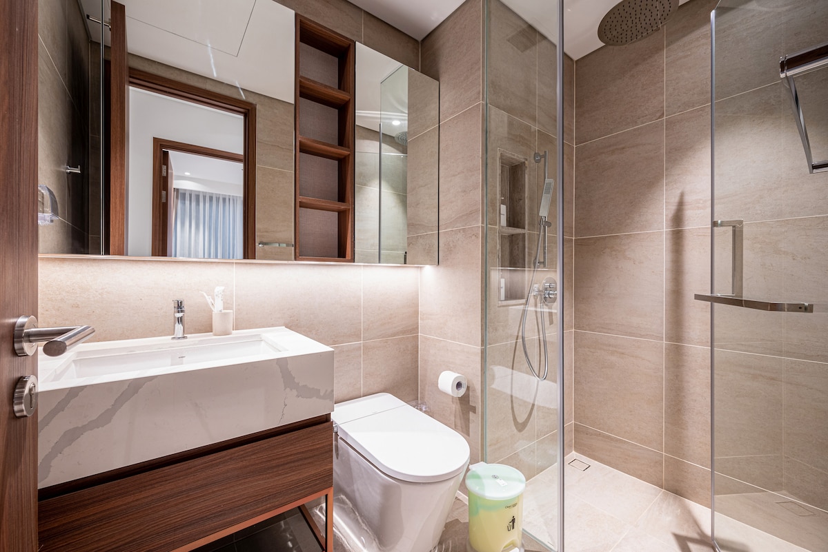 The modern bathroom features a sleek design with neutral-toned tiles. A spacious glass shower stall is visible alongside a contemporary sink and vanity. Shelving units are built into the wall, and a large mirror reflects soft illumination from the lighting above the sink.