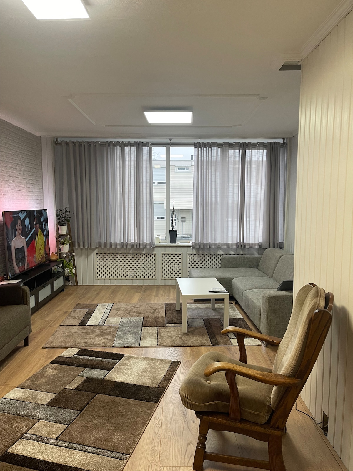 A spacious living area features a gray sofa, a light wooden coffee table, and a comfortable rocking chair. Natural light is welcomed through large windows adorned with sheer gray curtains. A patterned area rug adds texture to the floor, while a television is positioned nearby.