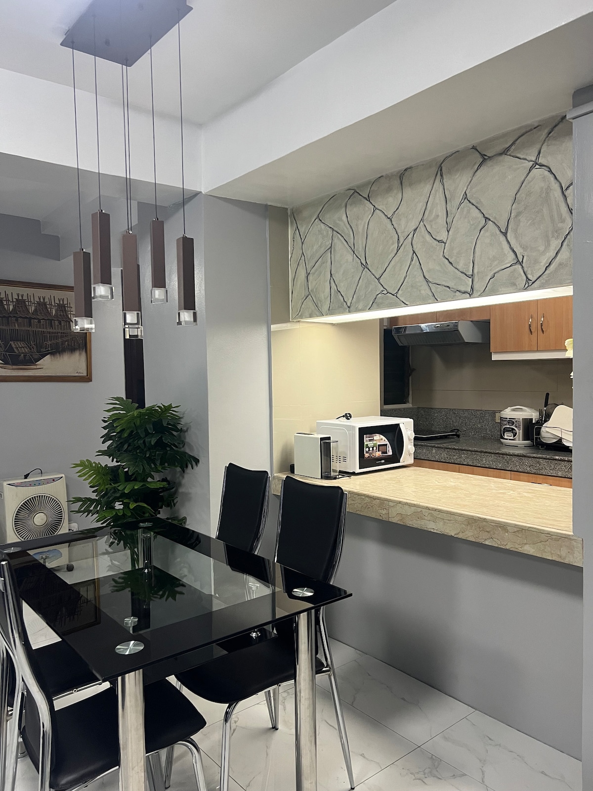 A modern dining area features a glass table surrounded by black chairs. A compact kitchen counter is visible, complemented by wooden cabinetry and essential appliances, including a microwave. Decorative plants add a touch of greenery to the space, enhancing its welcoming atmosphere.