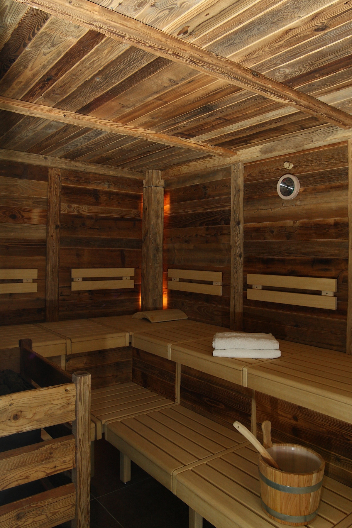 A wooden sauna is presented with multiple bench seating made of light-colored wood. The walls and ceilings consist of rustic wooden planks, creating a warm ambiance. A clock is mounted on one wall, and a wooden bucket and ladle are positioned on a bench, adding to the sauna's rustic charm.
